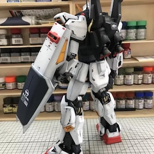 Op de afbeelding: Een gedetailleerd, geassembleerd model van een witte, zwarte en rode robot met een groot schild. De robot heeft ingewikkelde mechanische details, waaronder zichtbare gewrichten, panelen en stuwraketten. Het schild heeft tekst en een logo.