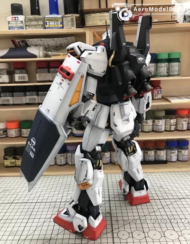 Op de afbeelding: Een gedetailleerd, geassembleerd model van een witte, zwarte en rode robot met een groot schild. De robot heeft ingewikkelde mechanische details, waaronder zichtbare gewrichten, panelen en stuwraketten. Het schild heeft tekst en een logo.