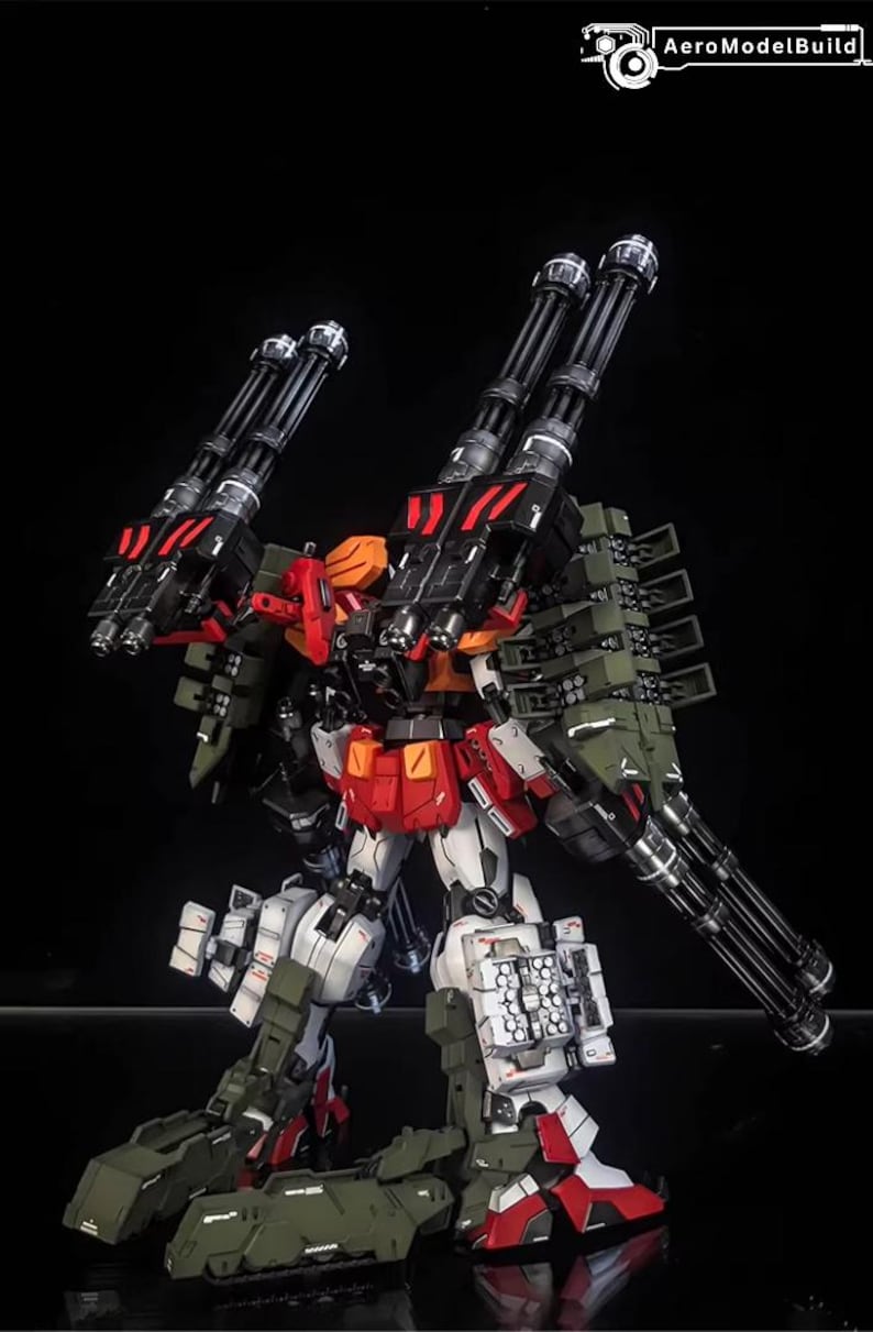 May include: A detailed model of a mech robot, mainly white, red, black, and olive green. The robot has multiple large cannons and missile launchers attached to its back and arms. The AeroModelBuild logo is in the top right corner.