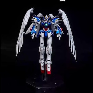 May include: A detailed model of a Gundam robot with large, white feathered wings. The robot features a color scheme of white, blue, red, and gold. It stands on a black stand against a black background. The text "AeroModelBuild" is in the top right corner.
