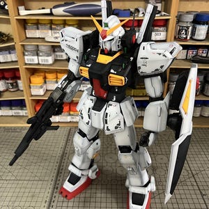 May include: A detailed, assembled Gundam model kit. The robot is primarily white with black, yellow, and red accents. It has a large shield, a rifle, and the text "AG004" on its shoulder. The model is posed on a cutting mat.