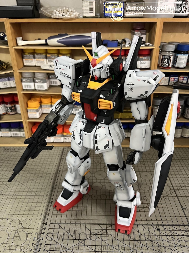 May include: A detailed, assembled Gundam model kit. The robot is primarily white with black, yellow, and red accents. It has a large shield, a rifle, and the text "AG004" on its shoulder. The model is posed on a cutting mat.