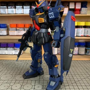 May include: A detailed model of a blue and black mecha robot with a shield and a rifle. The robot has yellow and red accents, with the number "03" visible. The shield has the word "TITANS" and a logo. The model is on a grid surface.