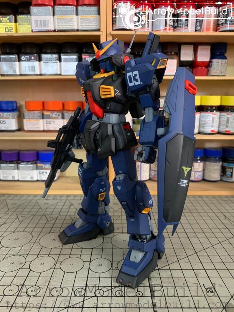May include: A detailed model of a blue and black mecha robot with a shield and a rifle. The robot has yellow and red accents, with the number "03" visible. The shield has the word "TITANS" and a logo. The model is on a grid surface.