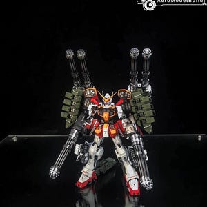 K&ouml;nnte beinhalten: Ein hochdetaillierter Gundam-Modellbausatz in Wei&szlig;, Rot und Orange mit umfangreicher Bewaffnung, darunter zwei gro&szlig;e, schultermontierte Kanonen und zwei handgef&uuml;hrte Gatling-Gesch&uuml;tze. Der Text AeroModelBuild ist oben zu sehen.