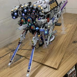 May include: A detailed model of a futuristic spacecraft, primarily white with accents of blue, gold, and black. The model features multiple light-up components, including blue and red lights, and is mounted on a clear stand. The text "ArrowModelBuild" is visible.