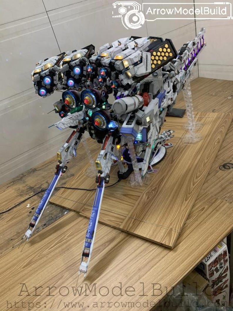 May include: A detailed model of a futuristic spacecraft, primarily white with accents of blue, gold, and black. The model features multiple light-up components, including blue and red lights, and is mounted on a clear stand. The text "ArrowModelBuild" is visible.