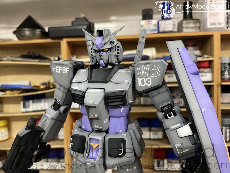 May include: A detailed model of a gray and purple mecha robot with the text "WB 103" and "EFSF" printed on the armor. The robot has a shield and is set against a blurred background of shelves.