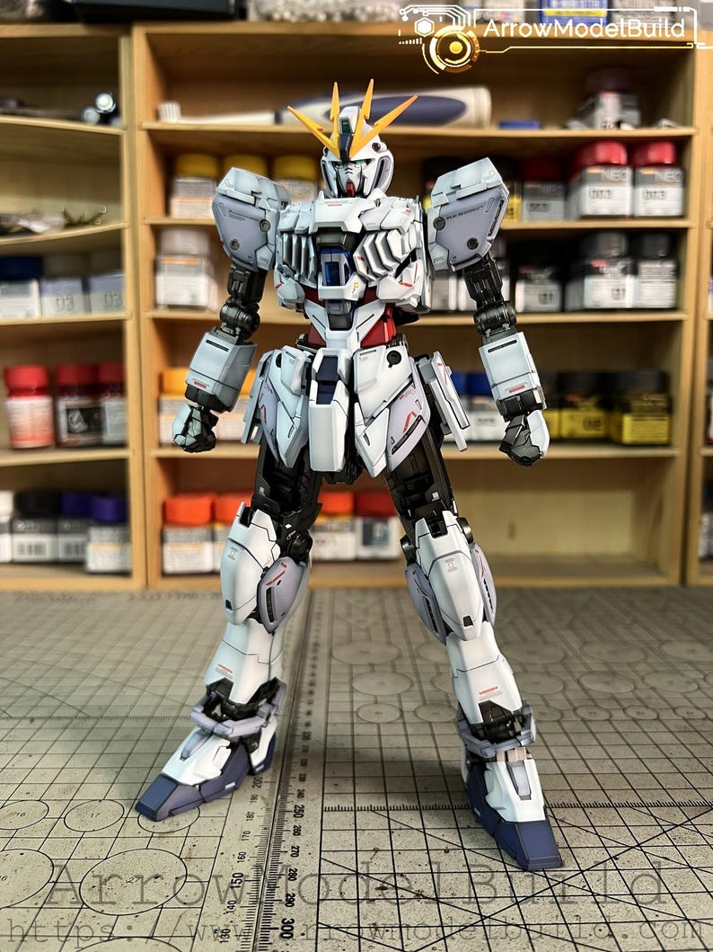 May include: A detailed model of a white, black, and blue robot with yellow accents. The robot is standing with its legs apart and arms at its sides. The model is on a cutting mat with measurements in inches. The text "ArrowModelBuild" is visible.