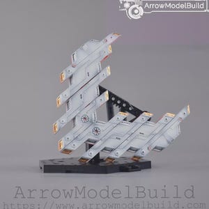 May include: A model kit of a futuristic spacecraft in white and grey, with gold and red accents. The model is mounted on a black base. The text "ArrowModelBuild" is visible.