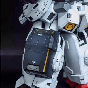 May include: Close-up of a detailed, articulated model robot. The model features a color scheme of white, gray, black, and gold accents. The robot's leg is visible, with a black shield and red foot. The model has visible text.