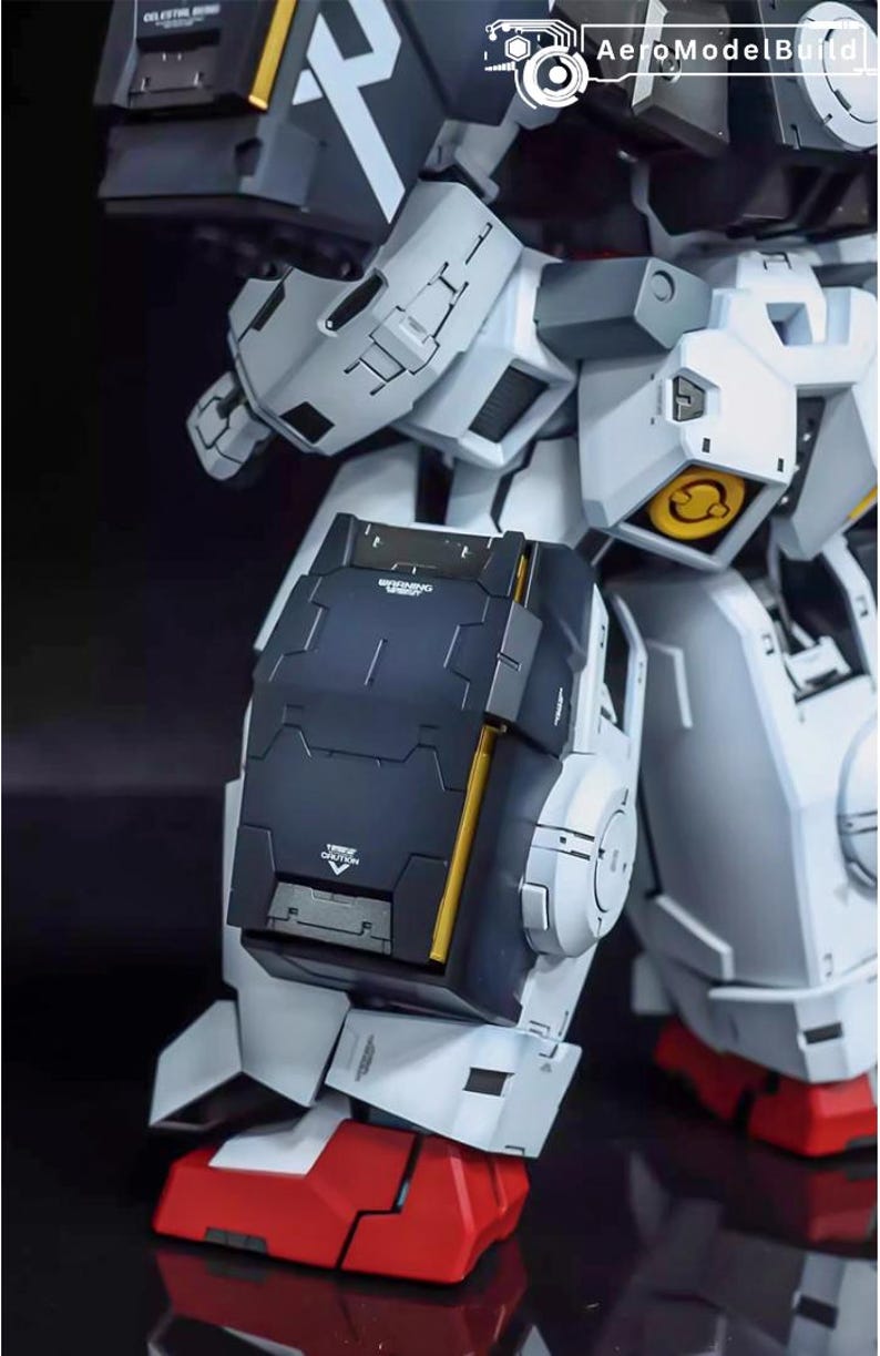 May include: Close-up of a detailed, articulated model robot. The model features a color scheme of white, gray, black, and gold accents. The robot's leg is visible, with a black shield and red foot. The model has visible text.