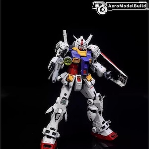 Op de afbeelding: Gedetailleerd model van een Gundam-robot, voornamelijk wit met rode, blauwe, gele en grijze accenten. De robot poseert met een schild en een wapen, tegen een zwarte achtergrond. De tekst "AeroModelBuild" staat rechtsboven.