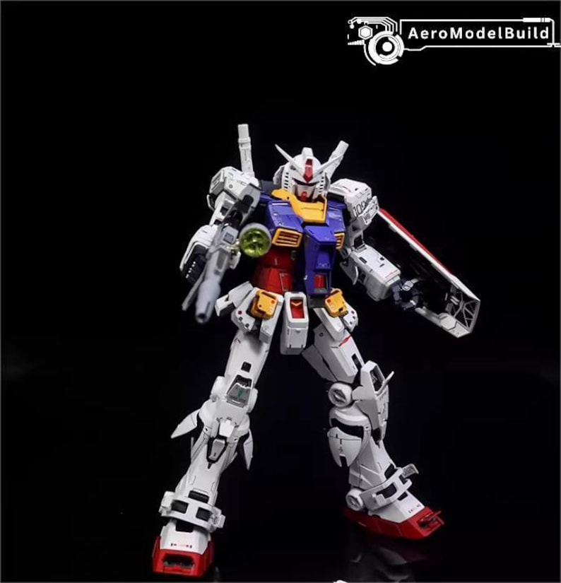 Op de afbeelding: Gedetailleerd model van een Gundam-robot, voornamelijk wit met rode, blauwe, gele en grijze accenten. De robot poseert met een schild en een wapen, tegen een zwarte achtergrond. De tekst "AeroModelBuild" staat rechtsboven.