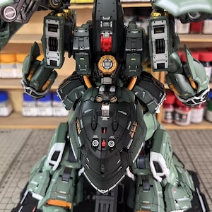 May include: A detailed, assembled model of a military-style robot, primarily in shades of green and black. The model features intricate mechanical details, including panels, joints, and a central core with gold and red accents. The model is on a black base.