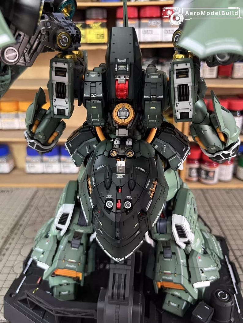 May include: A detailed, assembled model of a military-style robot, primarily in shades of green and black. The model features intricate mechanical details, including panels, joints, and a central core with gold and red accents. The model is on a black base.