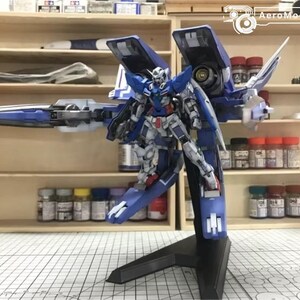 May include: A detailed model of a blue, white, and red robot with large wings and a long weapon. The model is mounted on a black stand. The background includes shelves with paint bottles and model-building supplies.