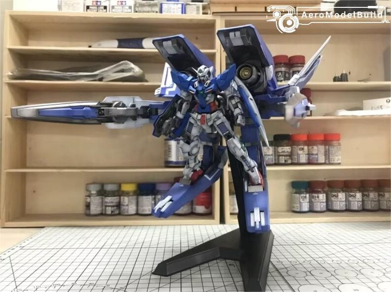 May include: A detailed model of a blue, white, and red robot with large wings and a long weapon. The model is mounted on a black stand. The background includes shelves with paint bottles and model-building supplies.
