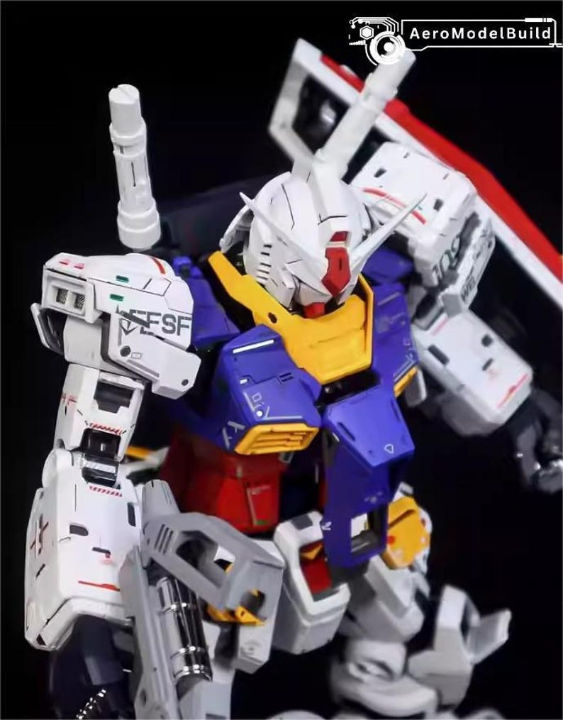 Op de afbeelding: Gedetailleerd model van een Gundam-robot, met een witte kop, rode en gele accenten, een blauwe en rode torso en grijze armen. Het model heeft ingewikkelde paneellijnen en markeringen, met de tekst "ESF" zichtbaar. Het AeroModelBuild-logo is zichtbaar.