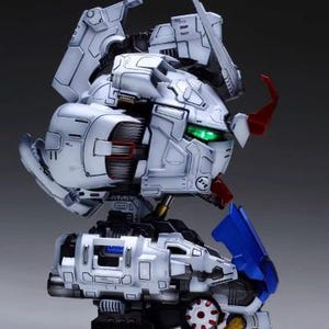 Może przedstawiać: Szczeg&oacute;łowy, biało-szary model głowy robota z czerwonymi, niebieskimi i złotymi akcentami. Model ma złożoną konstrukcję mechaniczną, z widocznymi kołami zębatymi i panelami. Oczy świecą na zielono. Widoczny jest tekst "ArrowModelBuild".