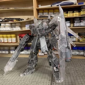 May include: A clear plastic model robot with articulated limbs and large weapons. The model is on a cutting mat, with shelves of paint and tools in the background. The model is made of clear plastic.