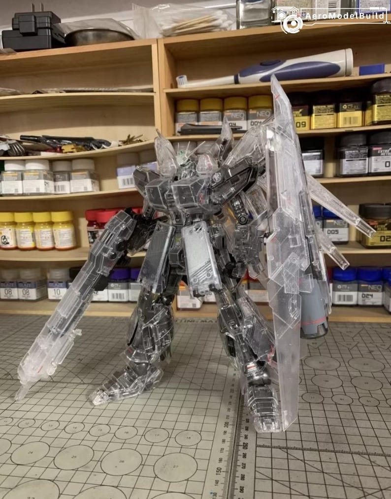 May include: A clear plastic model robot with articulated limbs and large weapons. The model is on a cutting mat, with shelves of paint and tools in the background. The model is made of clear plastic.