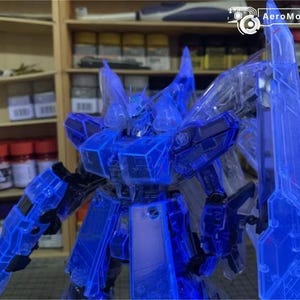 May include: A translucent blue model robot with sharp angles and a detailed design. The robot has large shoulder pads, a chest plate, and a large shield. The background includes shelves with various containers and the AeroModelBuild logo.