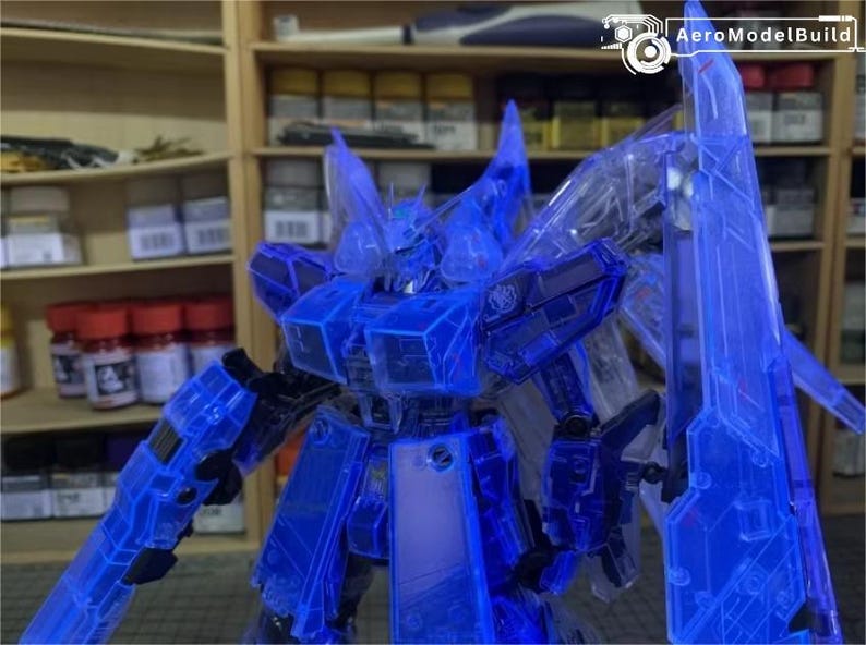May include: A translucent blue model robot with sharp angles and a detailed design. The robot has large shoulder pads, a chest plate, and a large shield. The background includes shelves with various containers and the AeroModelBuild logo.
