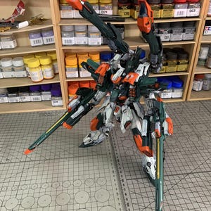 May include: A detailed model kit of a mech robot, featuring a color scheme of white, orange, and dark green. The model has intricate details and is posed on a cutting mat. Shelves filled with paint bottles are in the background.