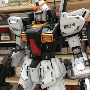 Op de afbeelding: Een gedetailleerd model van een Gundam-robot, voornamelijk wit met zwarte, rode, oranje en groene accenten. Het model heeft ingewikkelde paneellijnen en markeringen, waaronder "RX-178" en "SA6004". Het model staat tegen een achtergrond van planken met verfbenodigdheden.