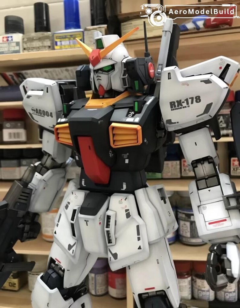 Op de afbeelding: Een gedetailleerd model van een Gundam-robot, voornamelijk wit met zwarte, rode, oranje en groene accenten. Het model heeft ingewikkelde paneellijnen en markeringen, waaronder "RX-178" en "SA6004". Het model staat tegen een achtergrond van planken met verfbenodigdheden.