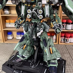 May include: A detailed model of a green and black robot with intricate mechanical details. The robot is posed on a black base, showcasing its articulated limbs and various components. The model features accents of yellow and white.