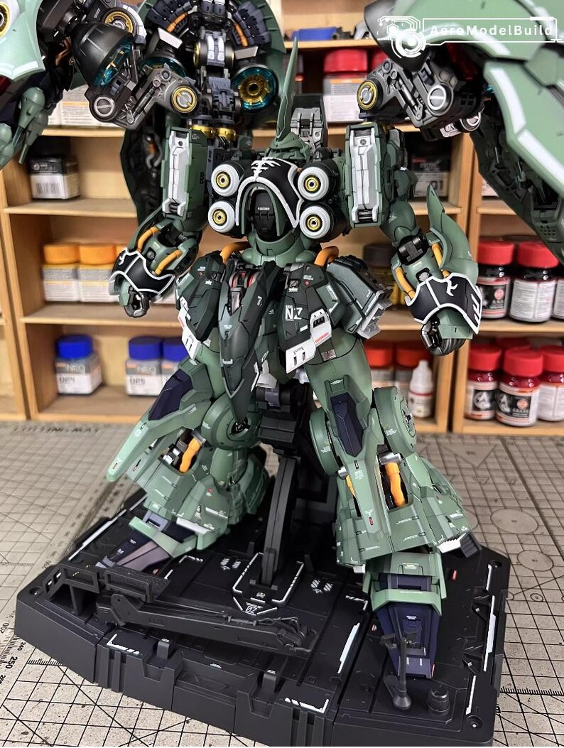 May include: A detailed model of a green and black robot with intricate mechanical details. The robot is posed on a black base, showcasing its articulated limbs and various components. The model features accents of yellow and white.