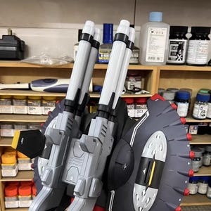 May include: AeroModelBuild model kit featuring a gray and white weapon system with two large barrels and a black shield with red accents. The model is set against a backdrop of paint supplies.