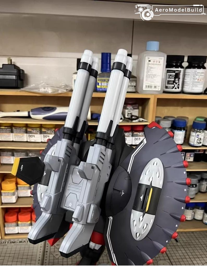 May include: AeroModelBuild model kit featuring a gray and white weapon system with two large barrels and a black shield with red accents. The model is set against a backdrop of paint supplies.