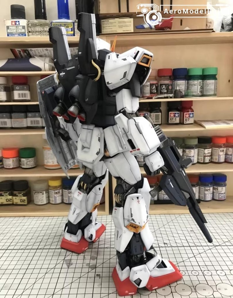 Op de afbeelding: Gedetailleerd model van een witte, zwarte en rode mecha-robot, met een geweer. De robot heeft een schild op zijn rug en staat op een oppervlak. Het model is zeer gedetailleerd met zichtbare paneellijnen en mechanische componenten.