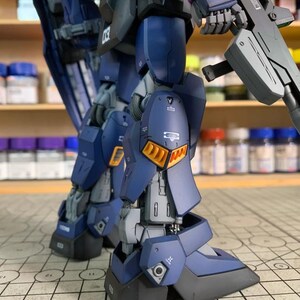 May include: A detailed, partially assembled model kit of a mech robot, primarily in shades of blue, gray, and black. The model features intricate panel lines, orange accents, and a weapon. The text "ArrowModelBuild" is visible.