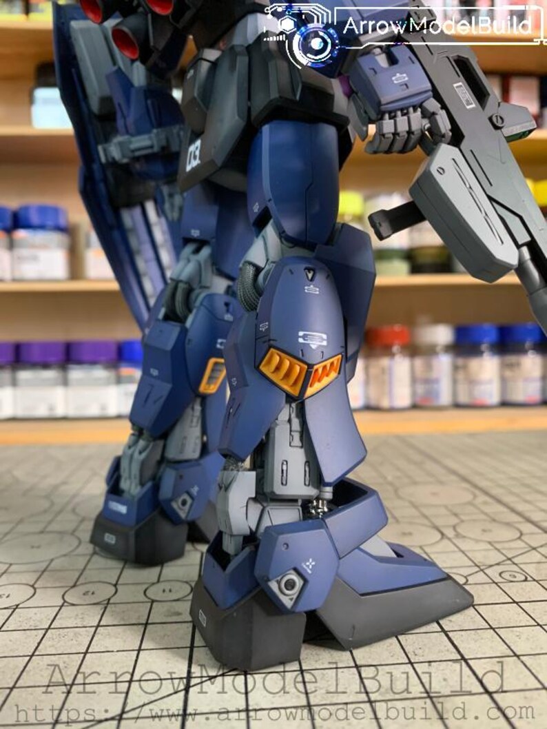 May include: A detailed, partially assembled model kit of a mech robot, primarily in shades of blue, gray, and black. The model features intricate panel lines, orange accents, and a weapon. The text "ArrowModelBuild" is visible.