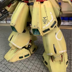 May include: Close-up of a yellow and white model robot's legs and lower torso. The model has a weathered appearance with black and brown accents. The word "TITANS" is visible on the leg.