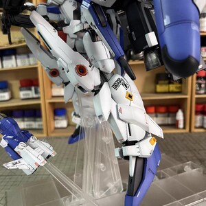 May include: A detailed model of a white, blue, and black mech robot with intricate designs. The model is partially assembled and features multiple panels, joints, and a small aircraft. The model is displayed on a clear stand, with the text "AeroModelBuild" visible.