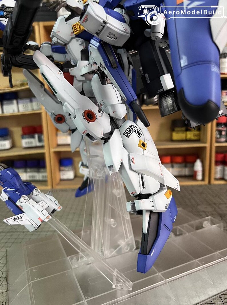 May include: A detailed model of a white, blue, and black mech robot with intricate designs. The model is partially assembled and features multiple panels, joints, and a small aircraft. The model is displayed on a clear stand, with the text "AeroModelBuild" visible.