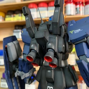 May include: A detailed model of a dark blue and black mecha robot with red accents on the thrusters. The model has the number "03" printed on the lower back. The text "TITANS ROYAL PROJECT" is visible on the shoulder.