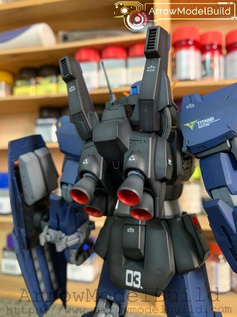 May include: A detailed model of a dark blue and black mecha robot with red accents on the thrusters. The model has the number "03" printed on the lower back. The text "TITANS ROYAL PROJECT" is visible on the shoulder.