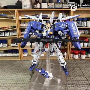 May include: A detailed model kit of a blue, white, and yellow mecha robot with a black rifle. The model is standing on a clear stand, with a smaller vehicle attached. The background shows shelves with paint bottles.