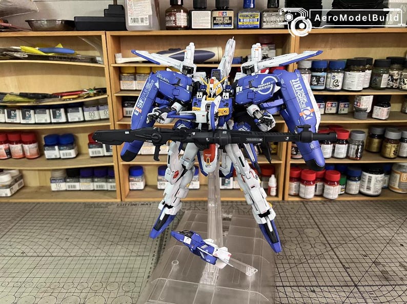 May include: A detailed model kit of a blue, white, and yellow mecha robot with a black rifle. The model is standing on a clear stand, with a smaller vehicle attached. The background shows shelves with paint bottles.