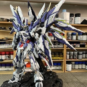 May include: A highly detailed robot model in white, purple, and blue, posed on a black base. The model features large, wing-like structures and intricate detailing. The background shows shelves filled with paint bottles, suggesting a hobbyist's workspace.