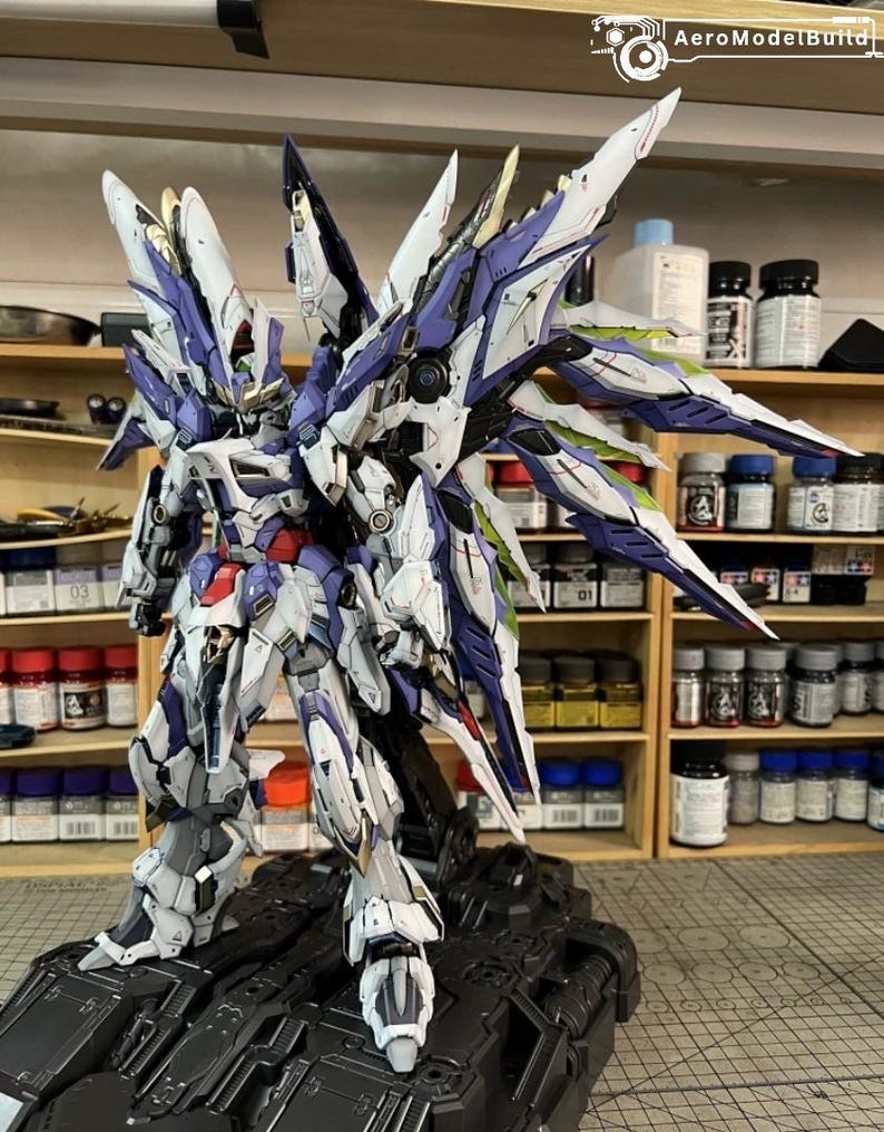 May include: A highly detailed robot model in white, purple, and blue, posed on a black base. The model features large, wing-like structures and intricate detailing. The background shows shelves filled with paint bottles, suggesting a hobbyist's workspace.