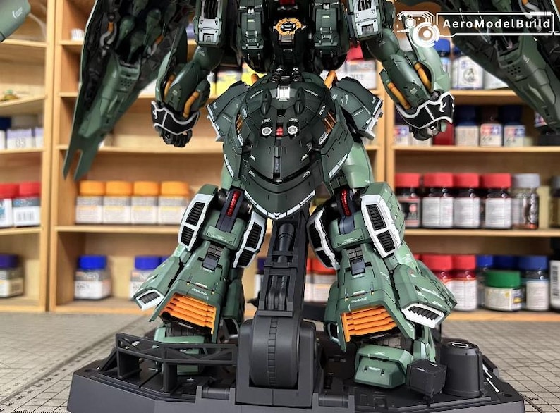 May include: A detailed, assembled model kit of a mech robot, predominantly in shades of green, black, and orange. The model is posed on a black stand, showcasing intricate mechanical details and panel lines. The AeroModelBuild logo is visible.
