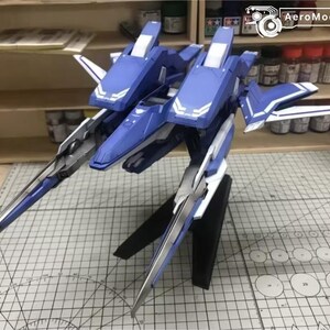 May include: AeroModelBuild model kit of a futuristic aircraft. The model is primarily blue and white, with silver accents. It features a dual-fuselage design and long, pointed wings. The model is displayed on a black stand.