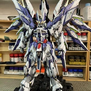 May include: A highly detailed model robot with large wings, predominantly white, blue, and grey, with red and gold highlights. The robot stands on a black base, displaying intricate mechanical details and a complex design, suitable for collectors.
